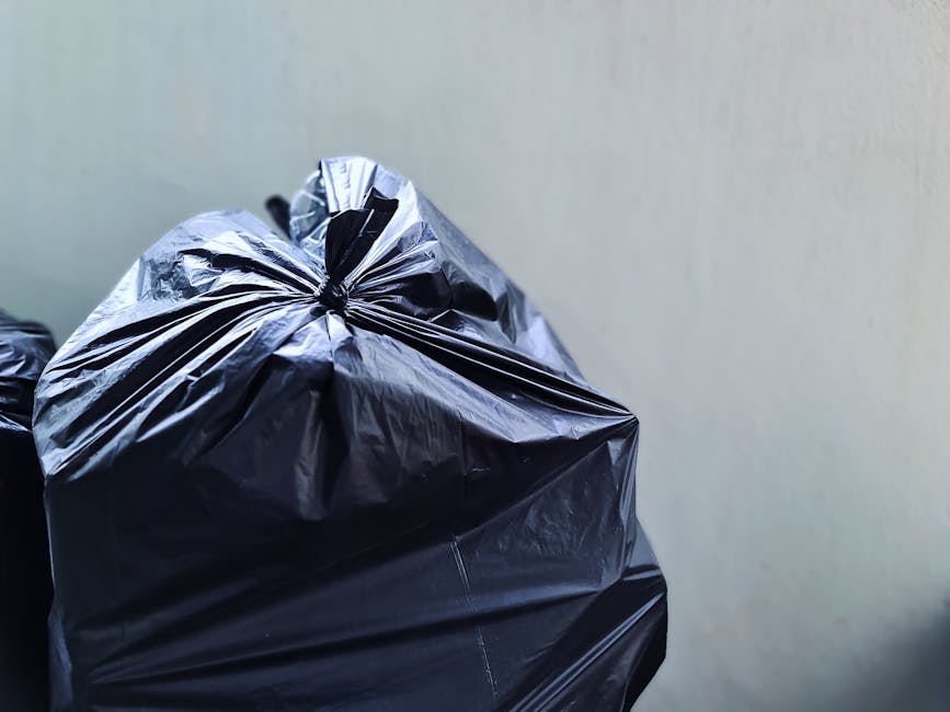 A close-up image showing a black plastic rubbish bag made from shiny, flexible material, tightly knotted at the top, positioned in the foreground against a plain, light-colored wall. The bag appears to be full and is set on a surface, likely on a driveway or pavement area outside. Additional black rubbish bags are partially visible behind it, suggesting a collection or removal process. The environment looks clean with natural lighting highlighting the reflective surface of the plastic, consistent with private rubbish disposal or alternative waste handling methods often carried out by waste management services like Rubbish Collection Tooting. The overall scene emphasizes the practical aspect of rubbish collection, suitable for professional waste removal from residential or commercial premises.