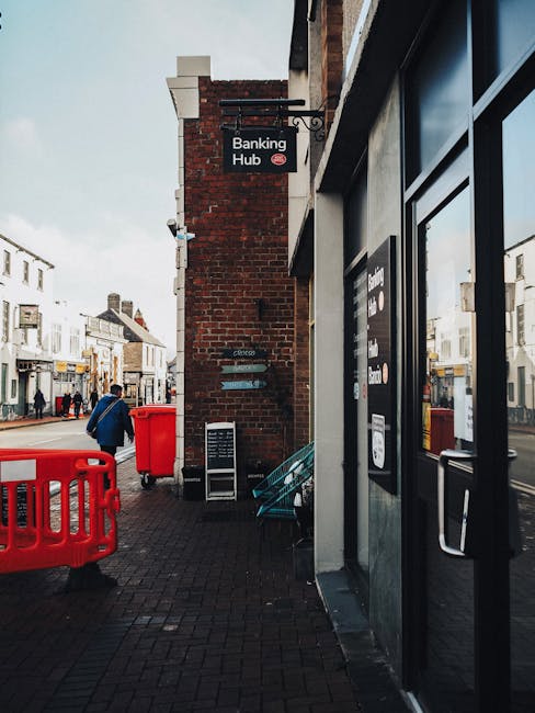 The image depicts a street corner with a brick building housing a banking hub, with a black and white sign hanging from the wall. In the foreground, there is a storefront with large glass windows and a door, which appears to be part of a commercial premises. The pavement is paved with dark bricks, and there are multiple red waste disposal items, including a large bin and a smaller bin, placed on the sidewalk. A person dressed in a blue jacket and black trousers is walking away from the camera, passing these waste bins. In the background, across the street, there are various shops and white terraced buildings with some visible signs and windows. The sky is overcast, providing diffuse daylight, and the surroundings suggest a busy urban area. The scene illustrates urban waste management practices, with external bins possibly used for private waste collection services, such as those offered by Rubbish Collection Tooting, in relation to alternative rubbish removal options on the high street.