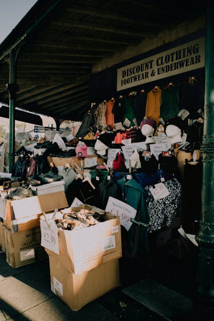 The image shows a street-side clothing and accessory stall under a covered pavement area, with a large black sign reading 'DISCOUNT CLOTHING FOOTWEAR & UNDERWEAR' mounted above. The stall displays a variety of second-hand or discounted items, including neatly arranged shoes, hats, and folded textiles on tables and shelves, along with hanging garments such as jackets, shirts, and sweaters in various colors and materials. Several cardboard boxes filled with additional goods are placed in the foreground, some with visible price tags indicating £2.99 or £3.99. The scene is outdoors, with shadows cast by sunlight, and the surrounding environment suggests an outdoor market setting, possibly part of a wider street market or informal retail area. This setup reflects a typical independent clothing stall that could be associated with alternative waste handling or second-hand goods collection, related to private waste disposal services like those offered by Rubbish Collection Tooting in the context of rubbish removal or clearance activities.