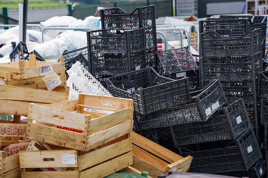 A large outdoor recycling or waste collection site displaying multiple black plastic crates and wooden boxes stacked and arranged in a disorderly manner. The black crates, made of sturdy perforated plastic, are placed in both upright and stacked positions, with some leaning against each other, revealing interior spaces filled with various waste or recyclable items. The wooden boxes are light-colored, unfinished, and rectangular, with some showing labels or tags attached. In the background, there are additional white bags and other waste containers, suggesting an active waste management area. The environment appears to be an open-air storage yard, possibly adjacent to a commercial or retail location, with natural daylight illuminating the scene. This snapshot highlights waste segregation or collection processes typical of private rubbish removal services, such as those offered by Rubbish Collection Tooting, for businesses requiring alternative waste handling solutions outside municipal services.